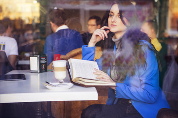 Brunette woman in business clothes: grey sweater and blue jacket sitting at the cafe near the window in european city drinking latte coffee and working using her tablet and smartphone. copy space