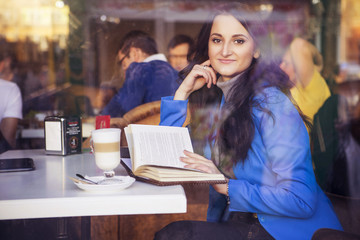 Brunette woman in business clothes: grey sweater and blue jacket sitting at the cafe near the window in european city drinking latte coffee and working using her tablet and smartphone. copy space