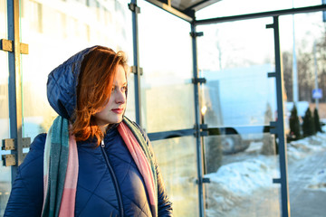 Young woman on a bus stop, caucasian girl with red hair waiting for the city bus, european female wearing hooded coat in the city bus station, sunset in the city