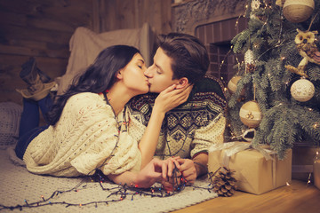 Beautiful brunette caucasian romantic loving couple in cozy warm sweaters in the cabin on a christmas new year eve, smiling, kissing, having fun with each other. Dim dark night light, toned image