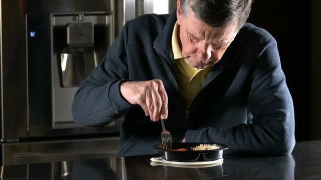 Lonely and depressed senior male sitting alone at kitchen table eating a microwaved ready meal of curry from plastic tray