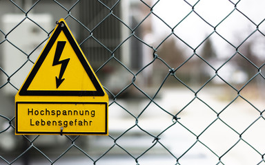 Schwarz-gelbes Warnschild an Maschendrahtzaun - Symbol für Lebensgefahr durch Stromschlag.