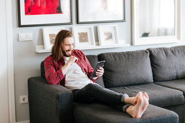 Cheerful bearded young man using tablet on sofa at home