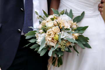 The groom in a suit and the bride in a white dress standing side by side and are holding bouquet of white and cream flowers.