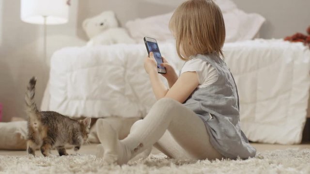 Cute Little Girl Sits on the Floor with Smartphone and Shoots Video of Her Striped Kitten Walking Around. Slow Motion. Shot on RED EPIC-W 8K Helium Cinema Camera.