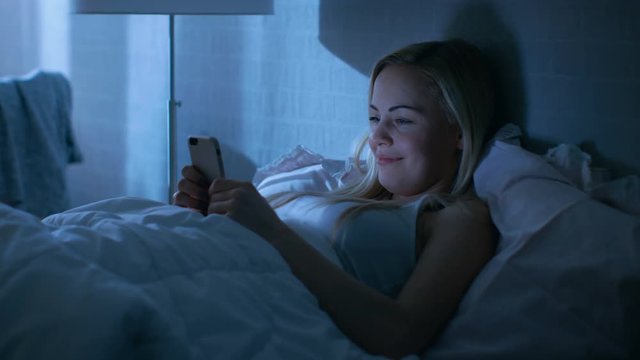 Beautiful Young Woman Lies in Bed at Night and Uses Smartphone.Shot on RED EPIC-W 8K Helium Cinema Camera.