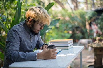 Young hipster beard man drinking coffee while reading books in home garden with nature. Education concept.