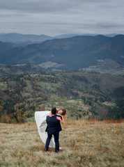 Naklejka na ściany i meble Groom in black suit hugs bride tender standing with her on hill with great mountain view