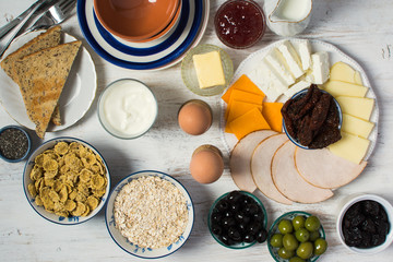 Healthy and various morning breakfast selection:  eggs, olives, cereals, ham, cheeses, bread, nuts on the white wooden table, selective focus. Top view