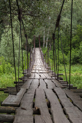 A suspended wooden bridge in green summer forest. Large cracks in the old boards and rusted cables of the suspension bridge. Forest landscape