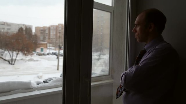 A man stands and thinks near window, he leaned against the wall, on the street cars ride
