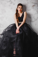 beautiful brunette girl in long black dress makeup red lips and manicure is sitting near the white gray wall looking at the camera