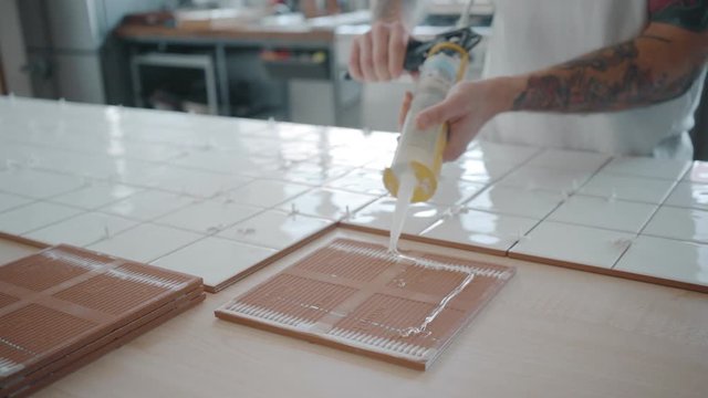 Tattooed hands using glue gun to apply transparent silicone adhesive on ceramic tile and put it on place close to other tiles on table, separated with white plastic spacers