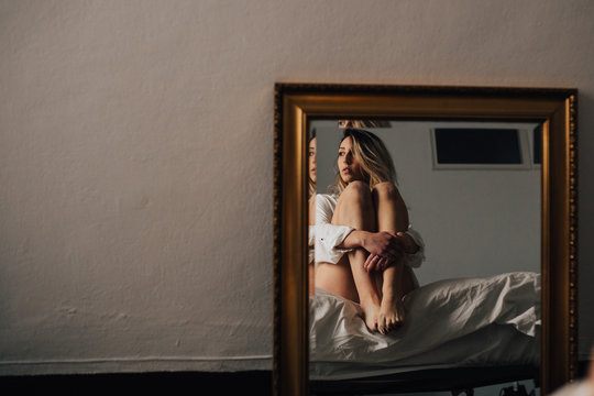 The reflection of female embracing her knees sitting on a bed. Horizontal indoors shot.