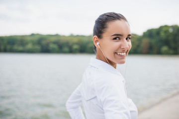 Portrait of young beautiful caucasian woman wearing sportswear looking at camera from back. Female athlete listening music with earphone. Smiling girl runner listen music in earphones from smartphone.