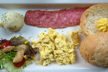 Breakfast consists of a slice of ham, bread, salad, potato salad ball and scrambled eggs on a white plate. Retro effect. Toned image.