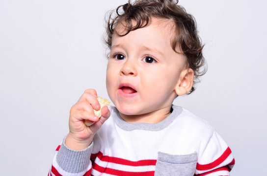 Portrait of a cute baby cringing while eating orange. One year old kid eating fruits by himself. Adorable curly hair boy being hungry biting on a slice of orange.