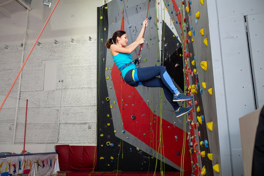 Happy sporty woman with climbing equipment hanging on a rope at indoor rock-climbing artificial wall