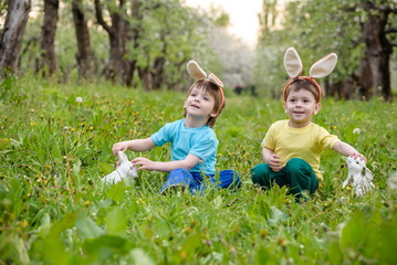 Naklejka na ściany i meble Two little kids boys and friends in Easter bunny ears during traditional egg hunt in spring garden, outdoors. Siblings having fun with finding colorful eggs. Old christian catholoc tradition