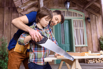 Low angle view of father teaching pensive son to saw wood in workshop