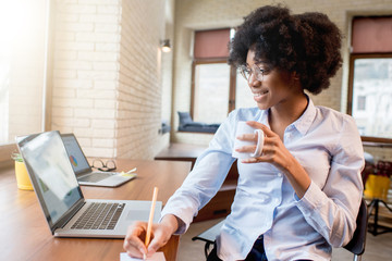 Young beautiful african businesswoman drinking coffee while sitting with laptop at the cafe or...