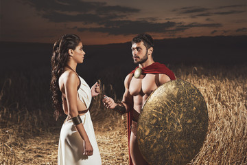 Woman wearing like greece giving amulet to man like spartan.