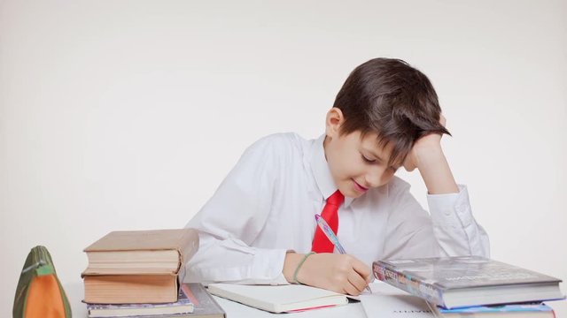 Young Caucasian kid school boy in red tie sitting at table learning writing surrounded by books on white background