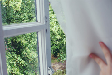 Mujer apartando una cortina para mirar por la ventana un parque cercano 