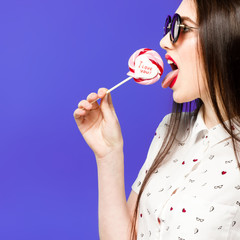 Young beautiful woman holding lollipop isolated on blue background. Happy girl wearing sunglasses eating multi colored candy. joyful and cheerful woman eating lollypop having fun