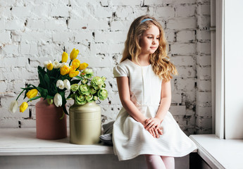 Little blonde girl in a white dress sitting by the window