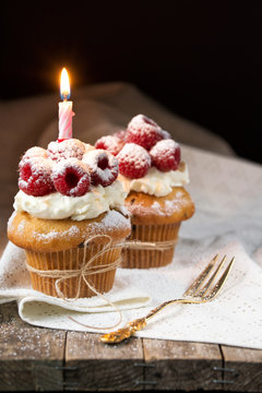 Muffins with soft cream and fresh raspberry powdered with sugar and decorated with burning candle, happy birthday card, rustic background