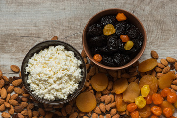 cottage cheese in white plate and the prunes in the brown plate. Prunes, dried apricots, dried mandarins and almonds on a light wooden background