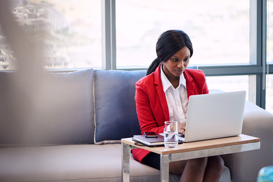 Attractive businesswoman wearing a red blazer busy working on her notebook computer in a business lounge, seated on a blue and grey couch with foreground blur visible in the image.