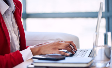 Generic image of a black woman's hand busy typing on a modern notebook with her head cropped out of the image while wearing a white shirt and red blazer as well as large windows in the background. - obrazy, fototapety, plakaty