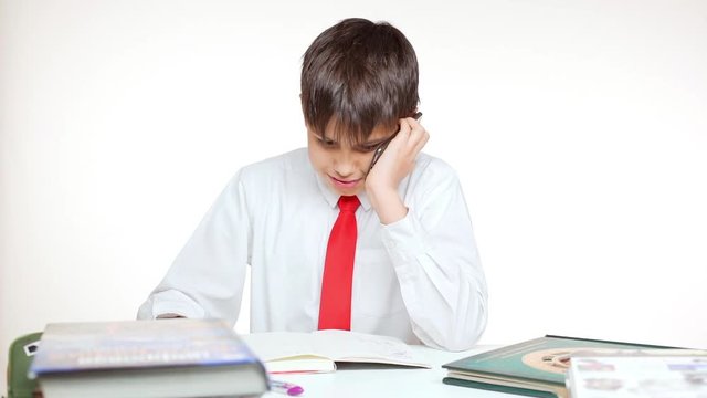 Young Caucasian school kid in red tie sitting at table writing talking on mobile phone on white background in slowmotion