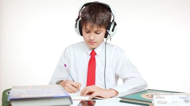 Young concentrated Caucasian school kid in red tie sitting at table wiritng listening music through headphones on white background in slowmotion