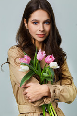 Fashion photo of a beautiful young woman with tulips in her hand .she dressed  in a beautiful brown trench coat, crimson dress.Spring concept. March 8. beautiful girl in stylish clothes.

