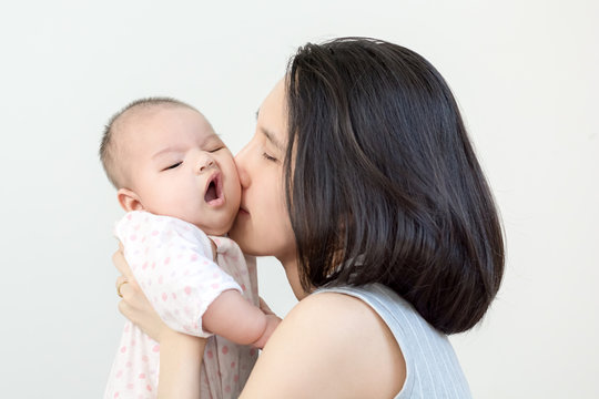 Portrait of happy asian mother kissing and hugging her cute little baby on grey background