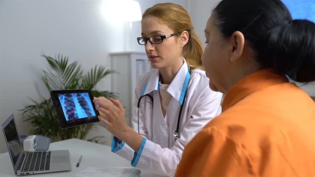 Attractive medical doctor is talking to female patient and showing her a X-ray picture on tablet while working in his office
