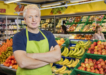 Smiling Salesman Standing Arms Crossed By Fruits In Store