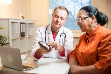 Smiling male doctor holding digital tablet, showing test results to patient in hospital. X-ray images on screen. Sick senior woman having a doctor appointment. Medical consultation.