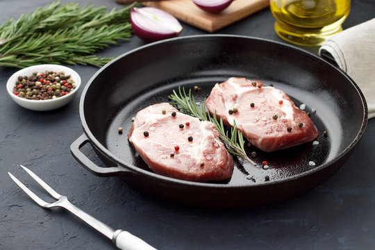 Two raw pork steak on frying pan with herbs and spices on dark stone background.