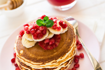 Traditional breakfast: stack of pancakes with banana slices and garnet seeds on white wooden table. Selective focus
