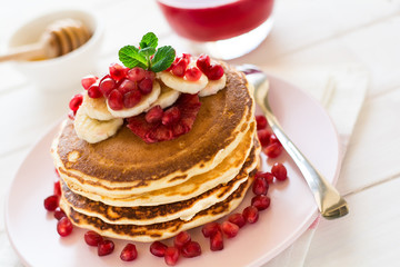 Traditional breakfast: stack of pancakes with banana slices and garnet seeds on white wooden table. Selective focus