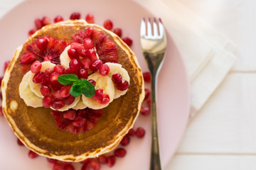 Traditional breakfast: stack of pancakes with banana slices and garnet seeds on white wooden table. Selective focus