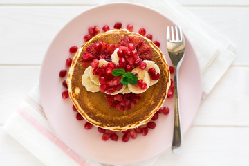 Traditional breakfast: stack of pancakes with banana slices and garnet seeds on white wooden table. Selective focus