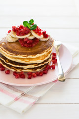 Traditional breakfast: stack of pancakes with banana slices and garnet seeds on white wooden table. Selective focus