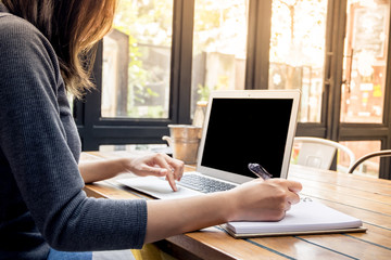 Beautiful young Asian girl working at a coffee shop with a laptop. Female freelancer writing on notebook in golden flare.