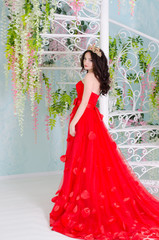 Beautiful woman in red long dress with a gold crown and earrings near forged spiral staircase