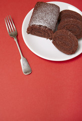 A sliced chocolate Swiss roll cake on a bright red background, with fork and empty space below